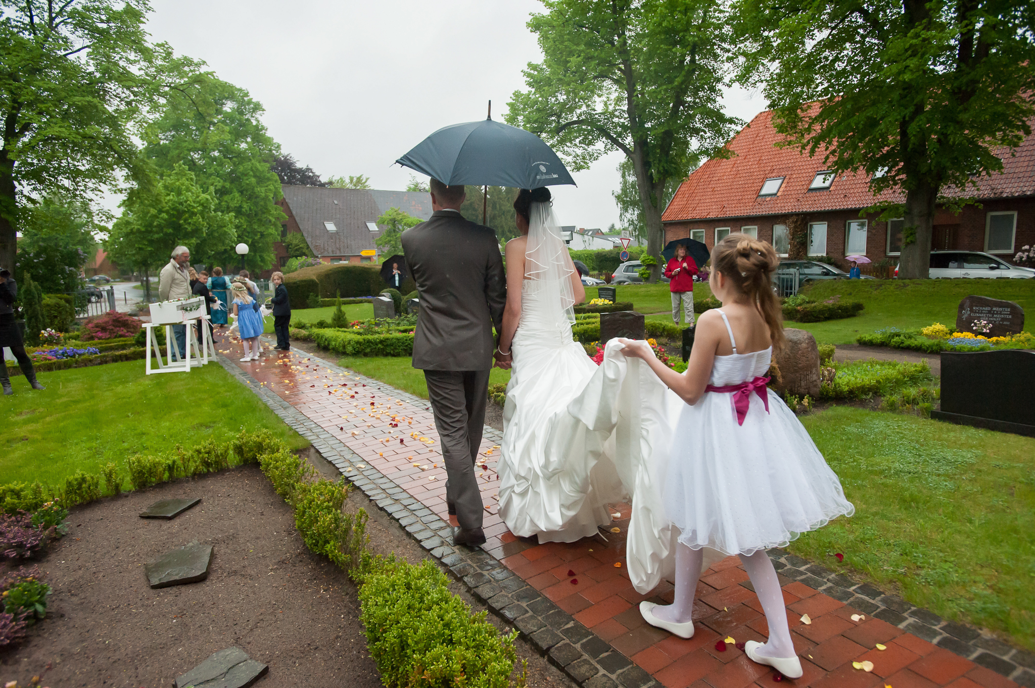 Brautpaar verlässt die Kirche bei regen, Blumenkind hält Brautkleid
